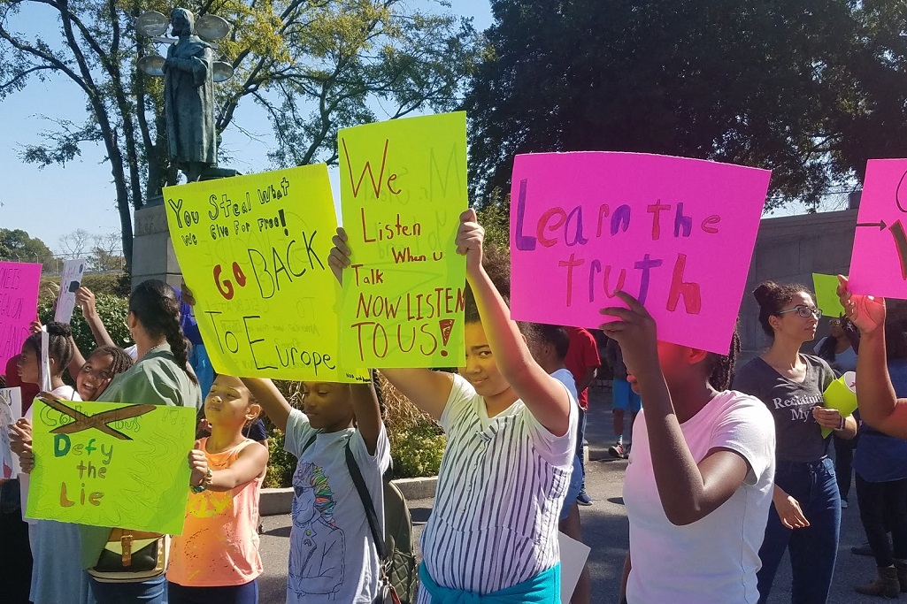“Defy The Lie”: Students and Native Americans Protest Columbus Statue